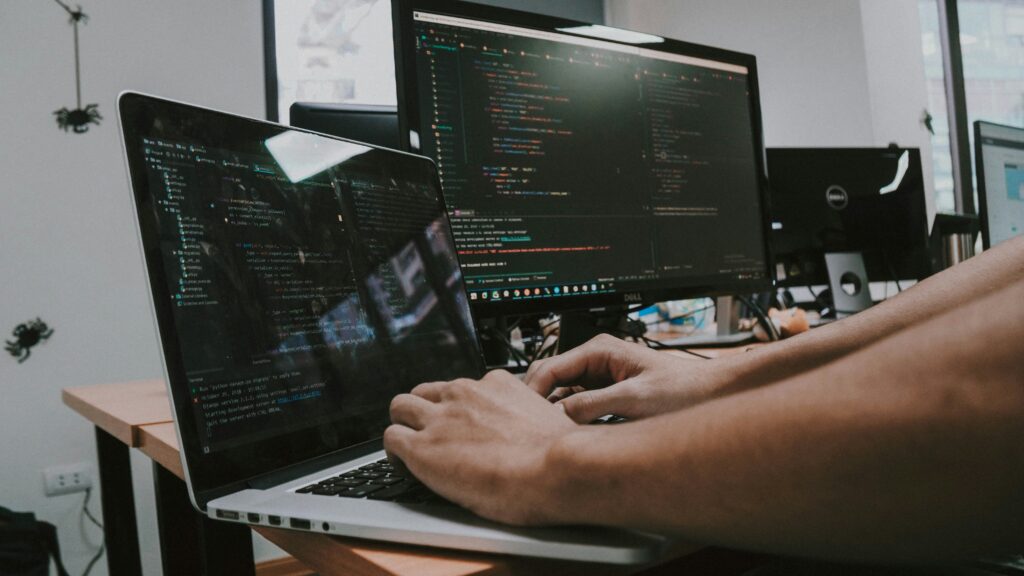 Male hands typing on a laptop that's placed next to a bigger desktop monitor